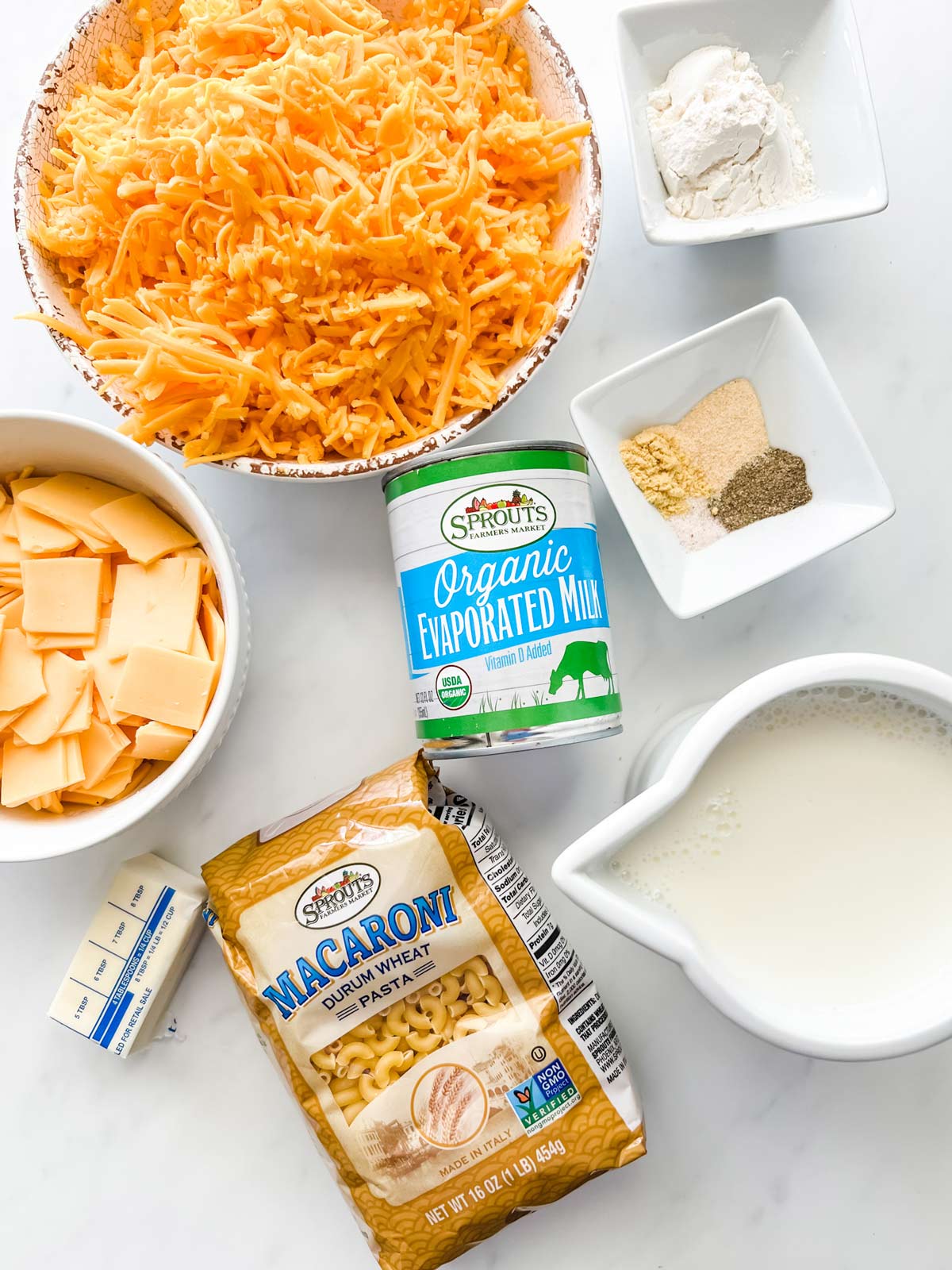 Overhead photo of shredded cheddar, flour, seasonings, american cheese, butter, macaroni noodles, evaporated milk, and milk.