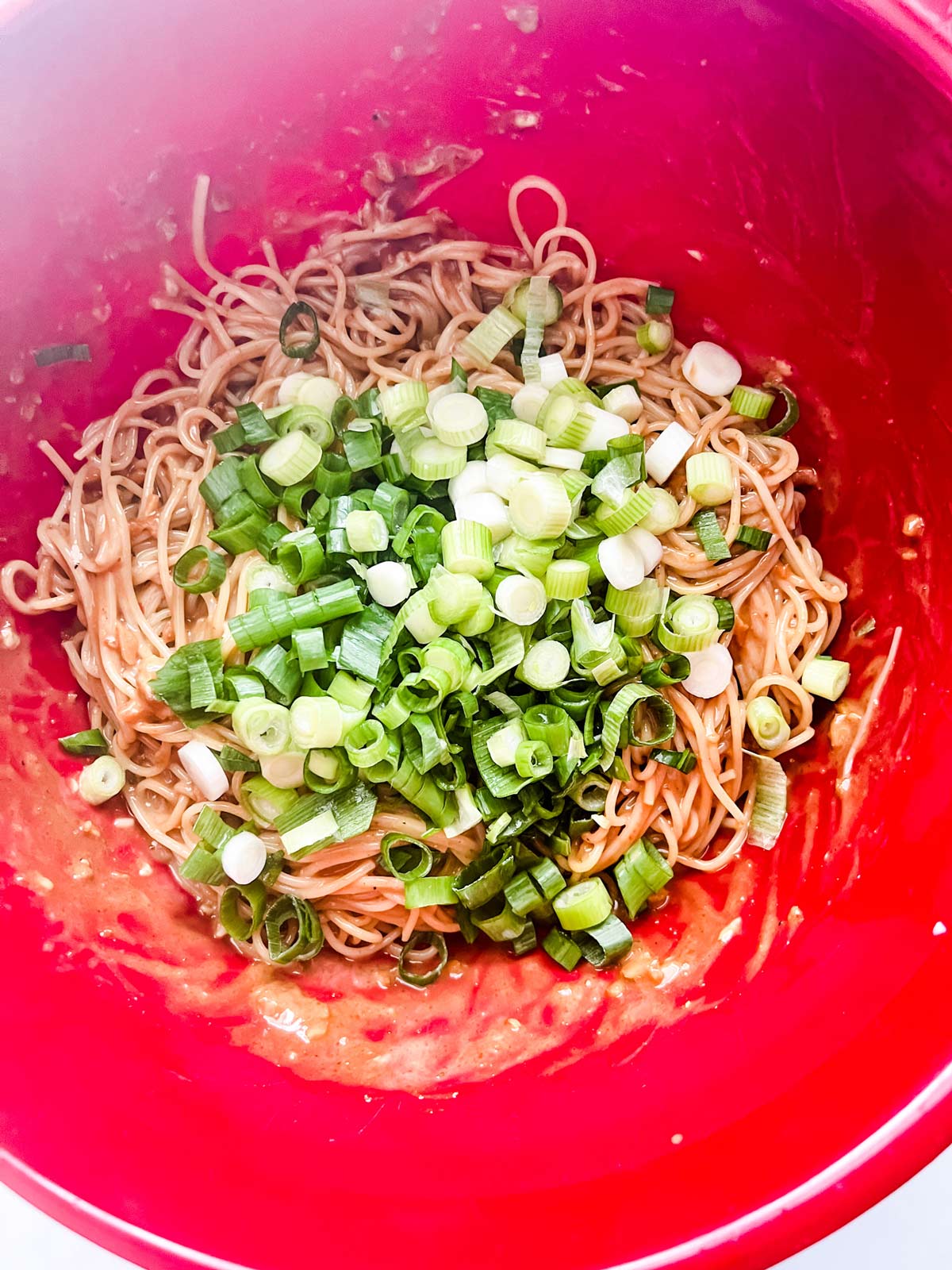 Cooked tahini pasta being tossed with chopped green onions in a red bowl.
