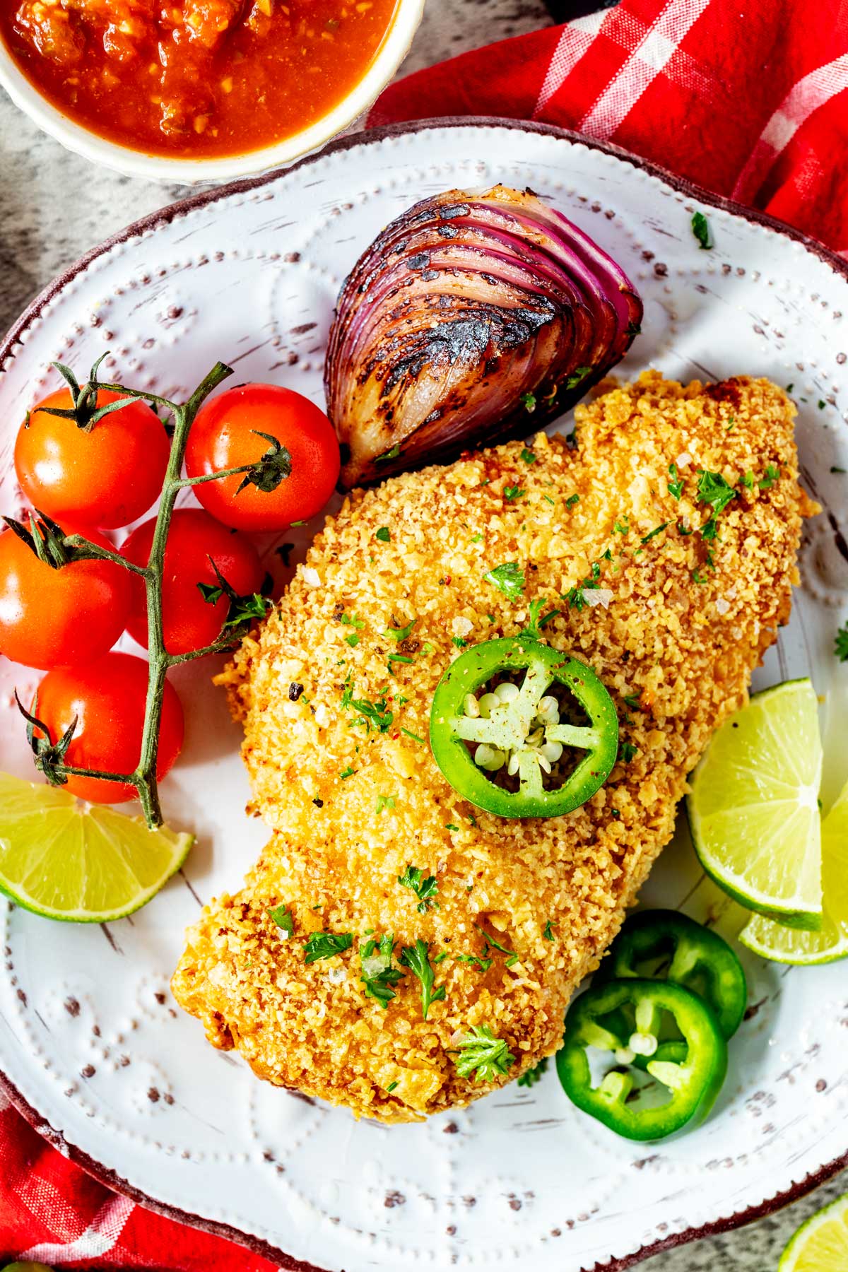 Overhead photo of a rustic plate with tortilla crusted chicken garnished with jalapeno, chopped cilantro, and lime.