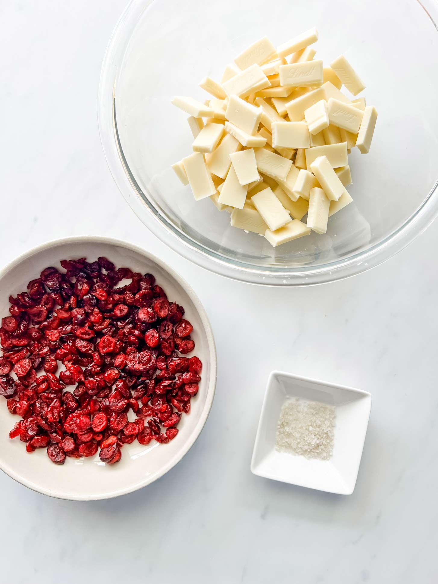 Overhead photo of a bowl of chopped white chocolate, a bowl of cranberries and a small dish of flakey salt.