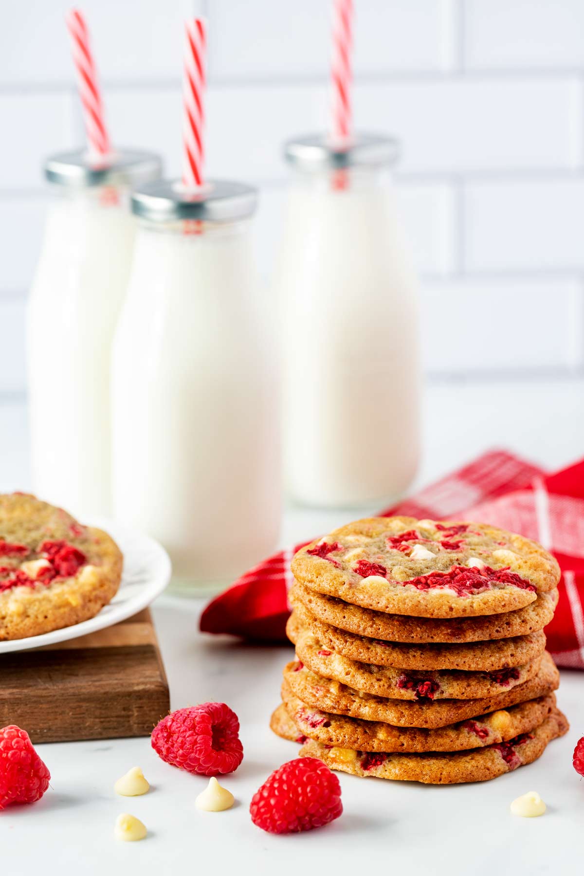 Side photo of a stack of white chocolate raspberry cookies with raspberries and chocolate chips beside it and jars of milk behind it.
