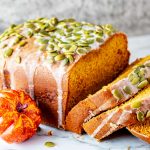 Square side photo of a partially sliced loaf of bread machine pumpkin bread with a small pumpkin and pumpkin seeds next to it.