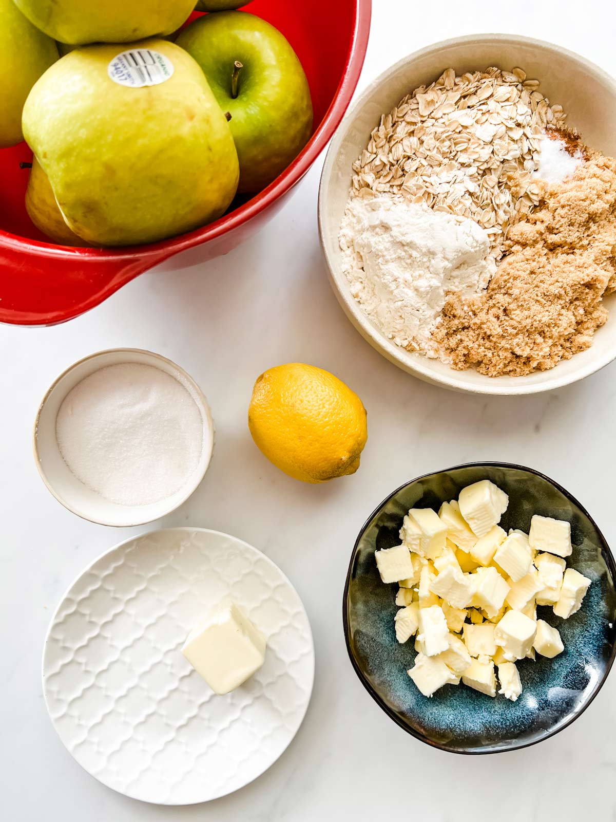 Overhead photo of apples, flour, oats, brown sugar, butter, lemon, and white sugar in prep bowls.