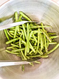 Photo of green beans in a bowl being tossed with oil, lemon juice and seasonings with tongs.