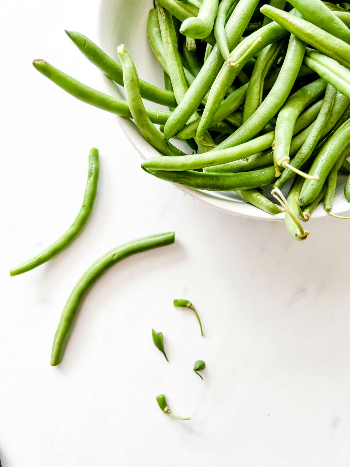 A bowl of green beans next to two green beans that have been trimmed.