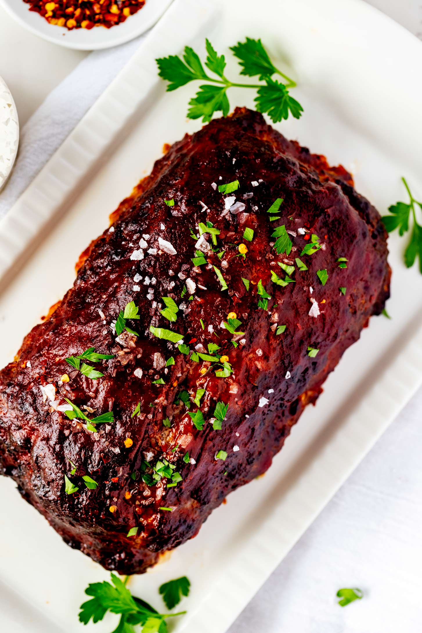 Overhead photo of meatloaf on a rectangular white platter garnished with flakey salt and parsley.