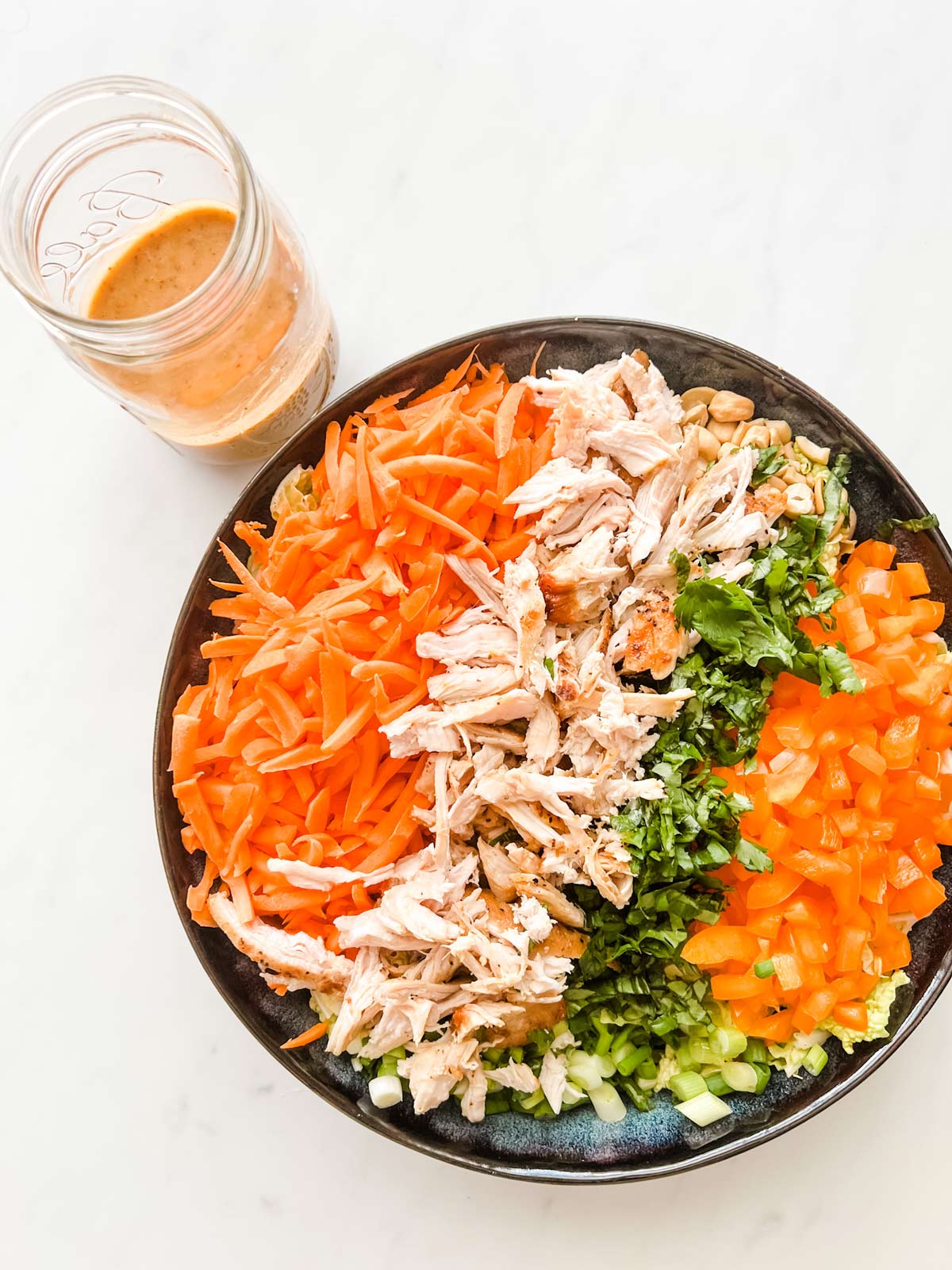 A large navy blue bowl with shredded napa cabbage, shredded carrots, cooked shredded chicken, cilantro, basil, and diced pepper sitting next to a jar of thai salad dressing.