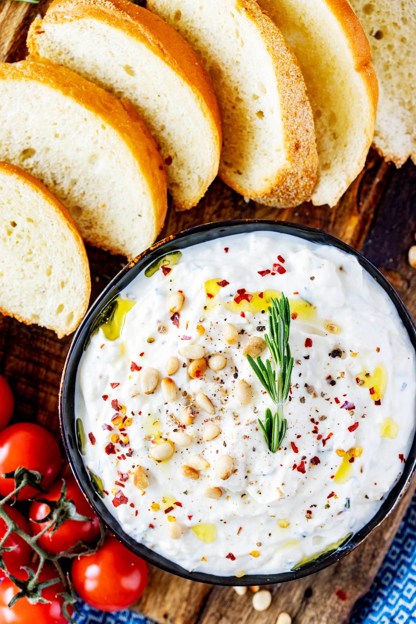 Overhead photo of a small bowl with whipped ricotta dip garnished with pine nuts and crushed red pepper flakes and surrounded by baguette slices.
