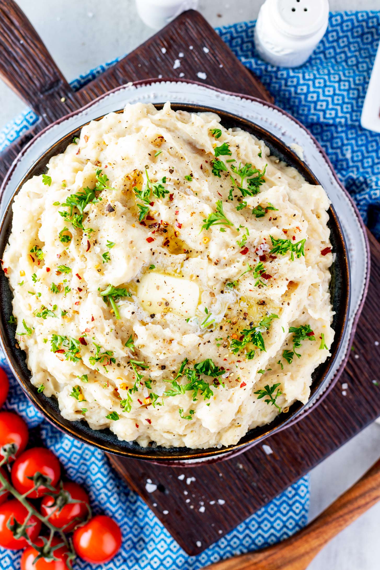 Overhead photo of a bowl of mashed potatoes garnished with parsley, flaky salt,and crushed red pepper flakes.
