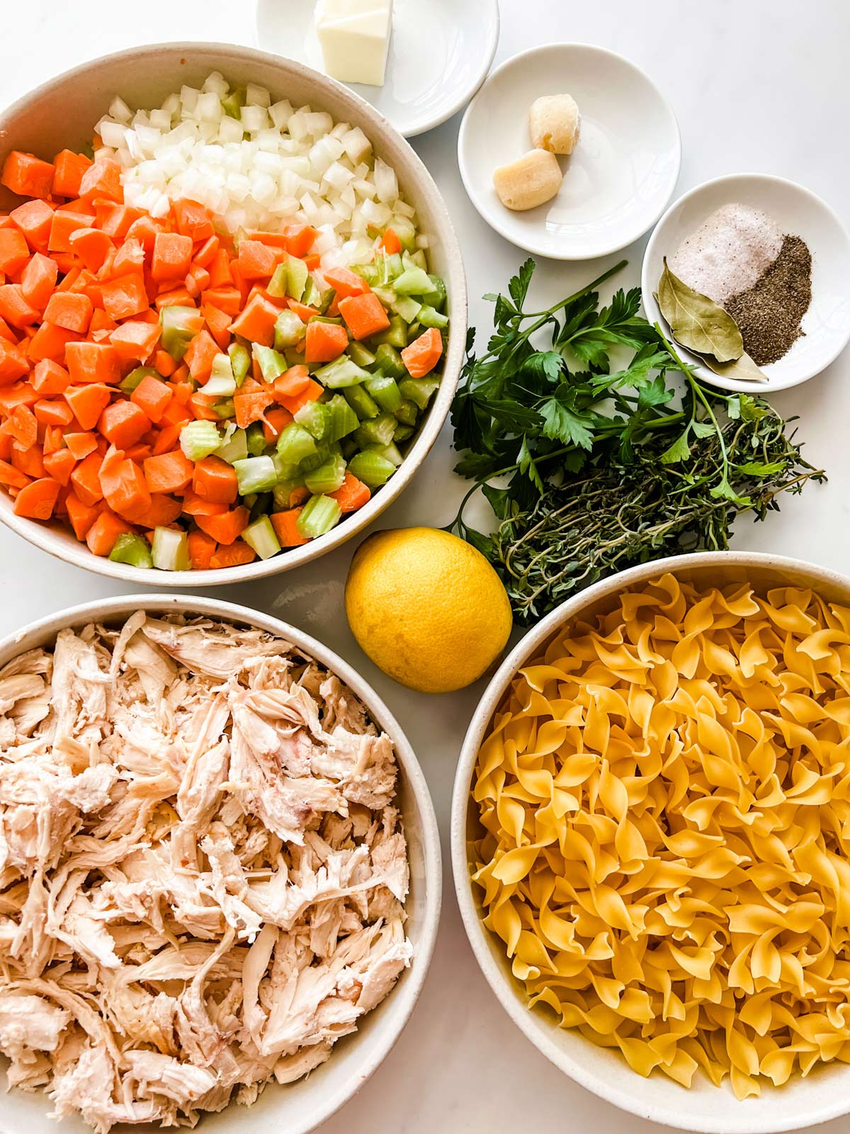Photo of butter, garlic, seasonings, fresh herbs, lemon, pasta, shredded chicken, carrot, celery, and onion in prep bowls.