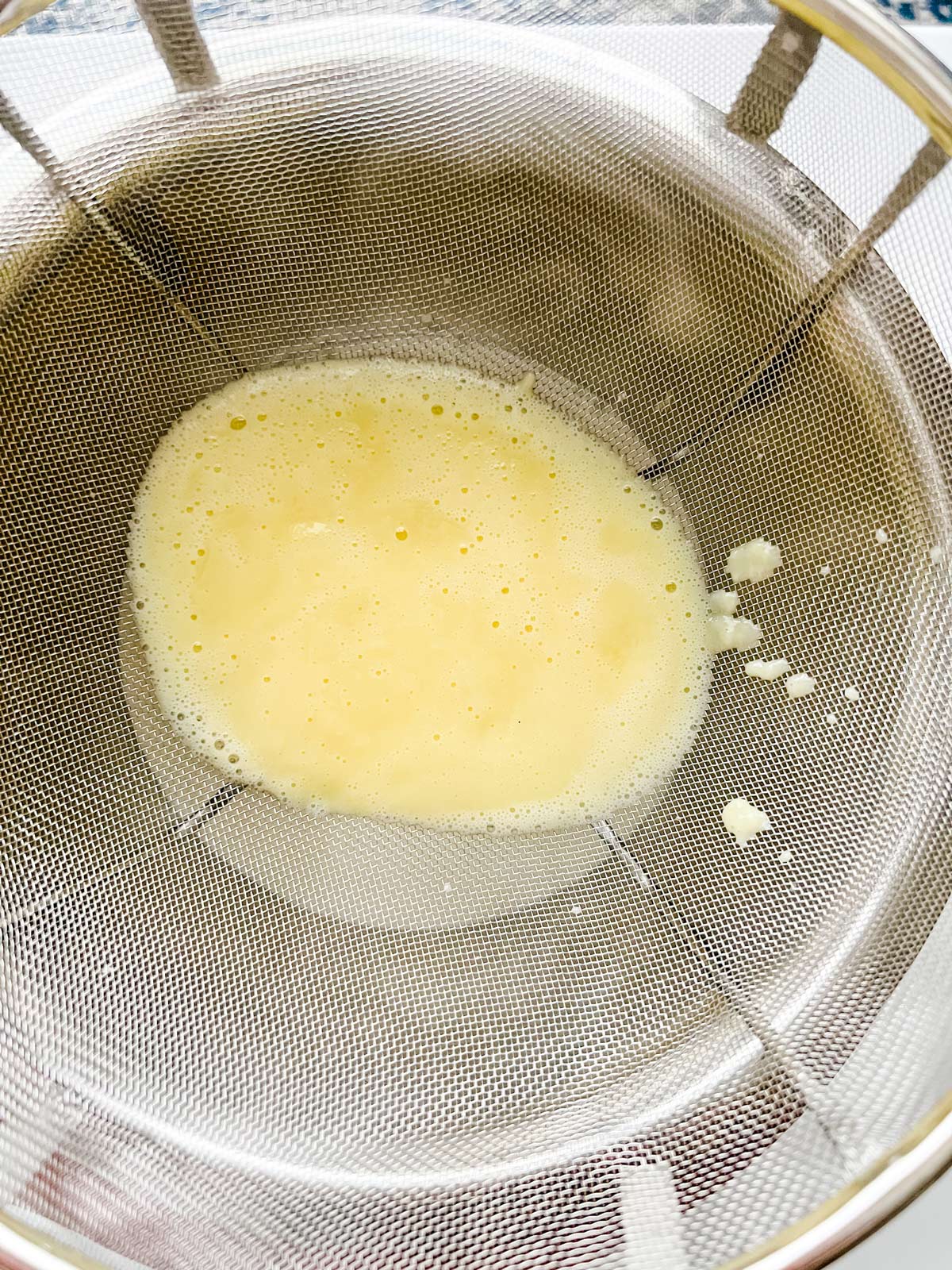 A cooked egg and milk mixture being strained into a bowl of heavy cream.