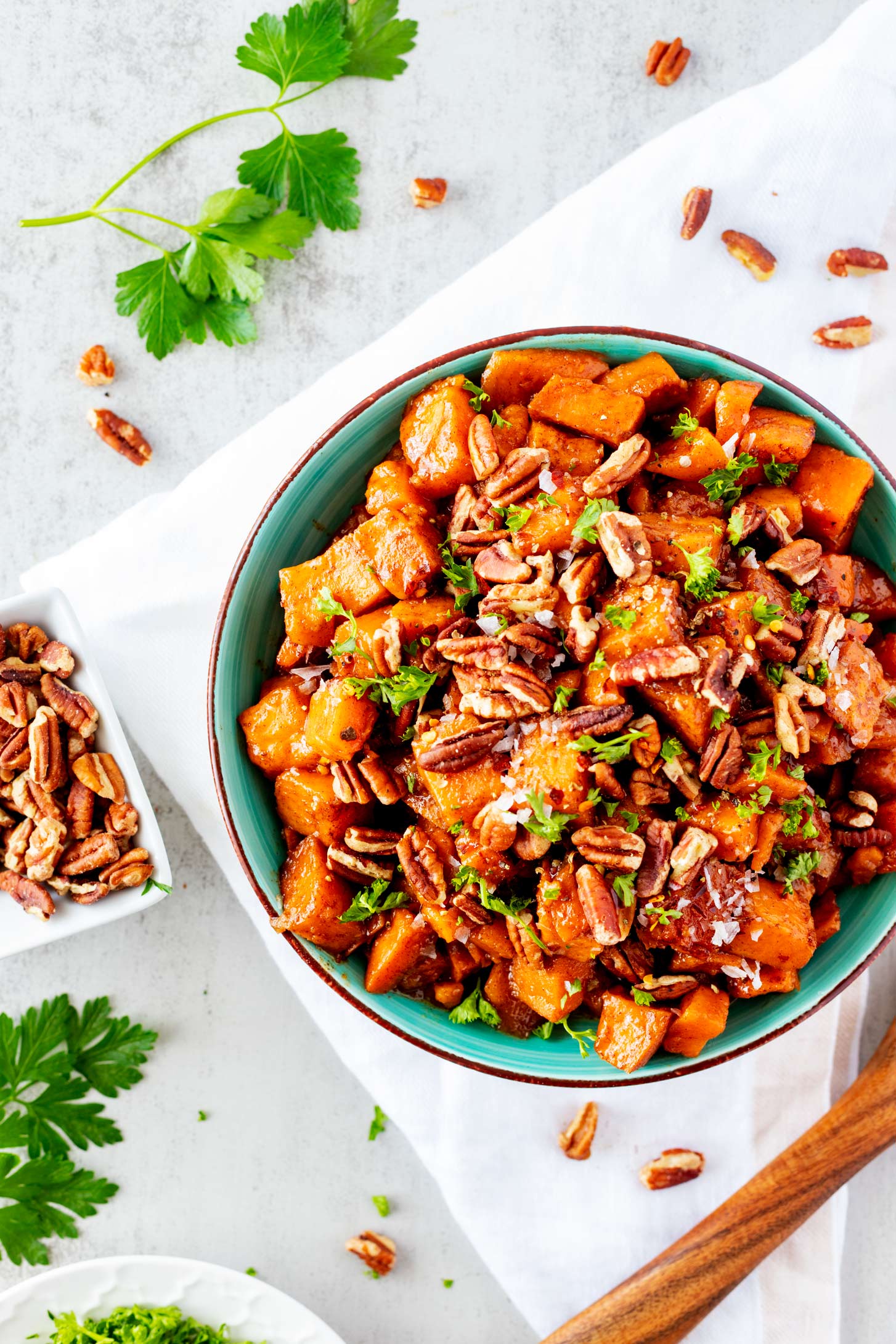 Overhead photo of a teal bowl of slow cooker sweet potatoes sitting on a white napkin and garnished with parsley and pecans.
