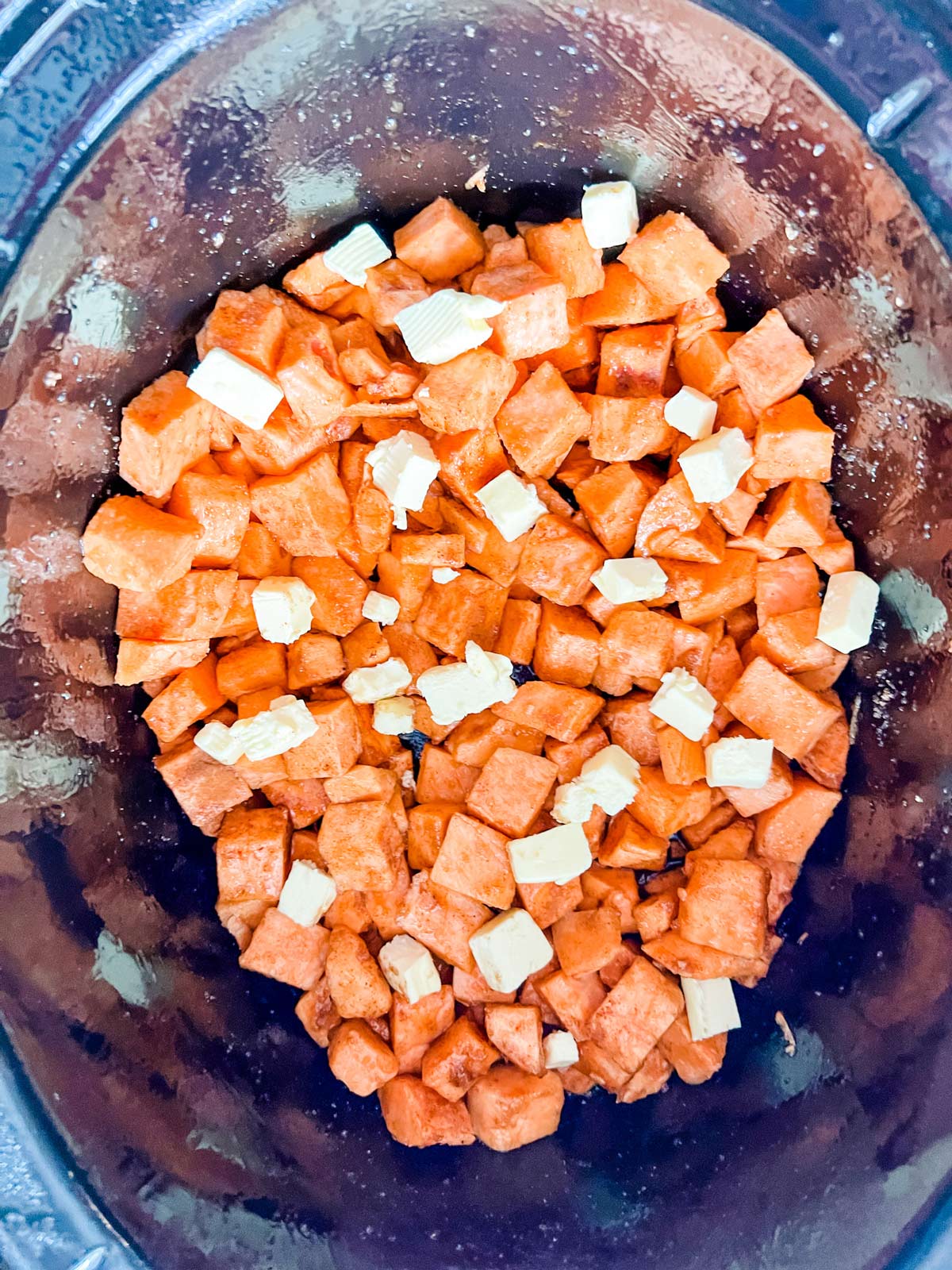 Sweet potatoes tossed with maple syrup, sugar, and seasonings and dotted with butter in a slow cooker ready for cooking.