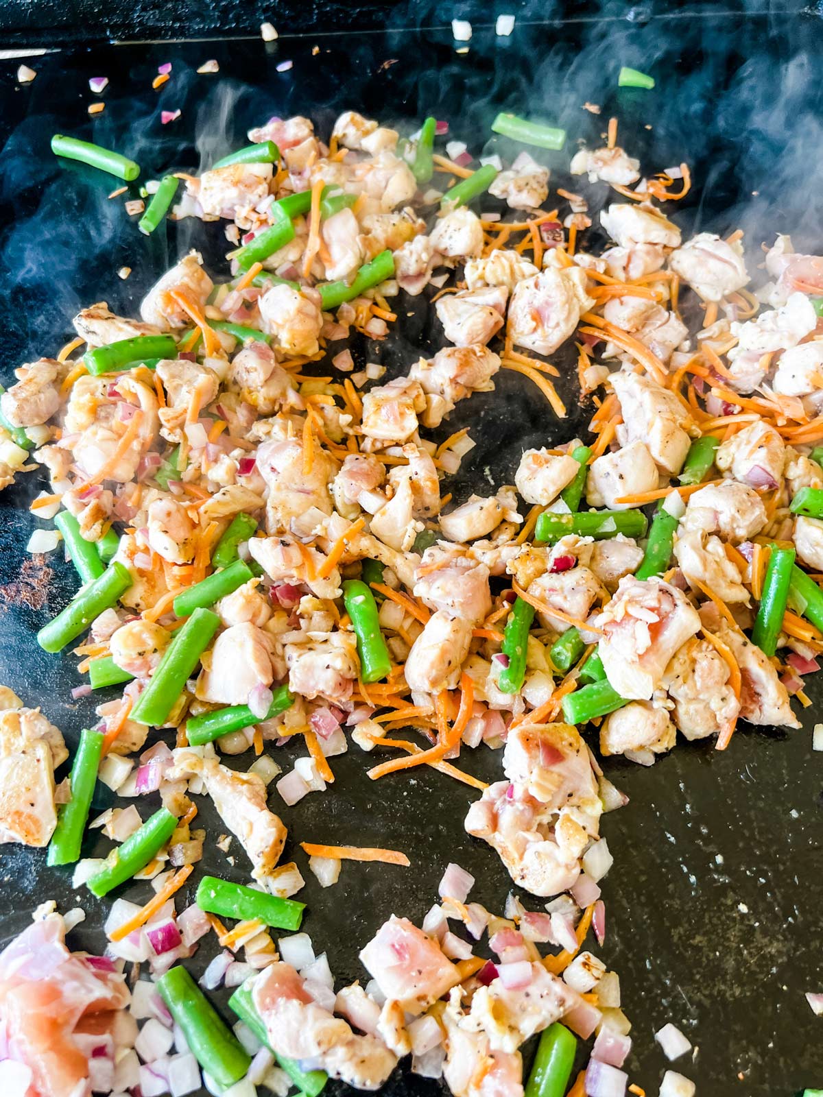 Chicken, red onion, carrot, and green beans cooking on a Blackstone griddle.