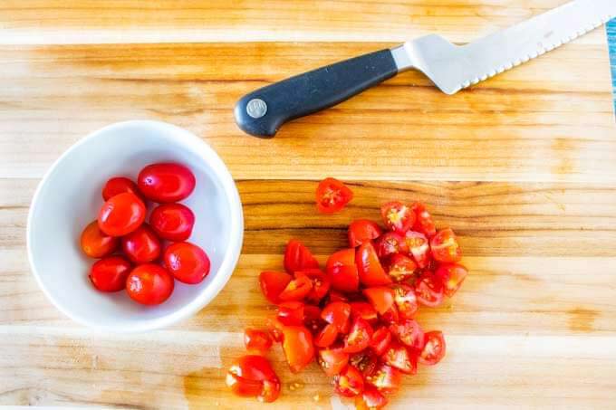 A bowl of grape tomatoes sitting on a cutting board with a knife and chopped tomatoes.
