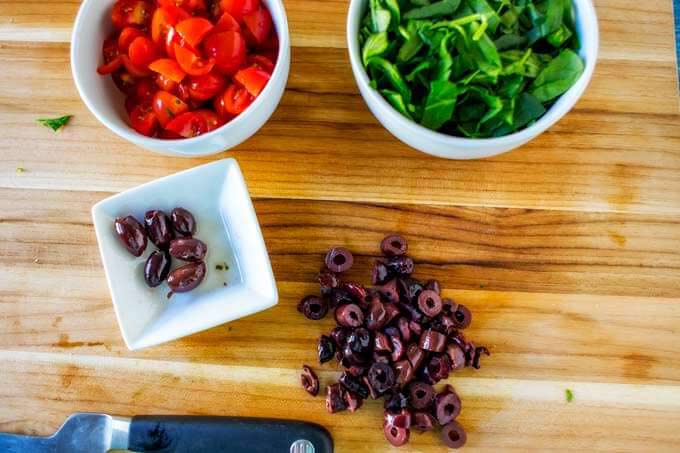 A cutting board with olives being sliced and a bowl of chopped tomato and chopped spinach.