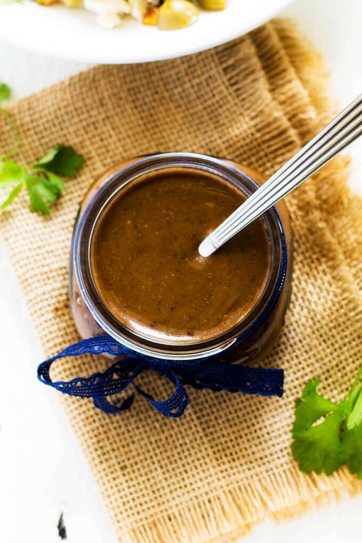 Photo of a Balsamic Vinaigrette Dressing Recipe in a glass jar with a blue ribbon sitting on a rustic fabric.