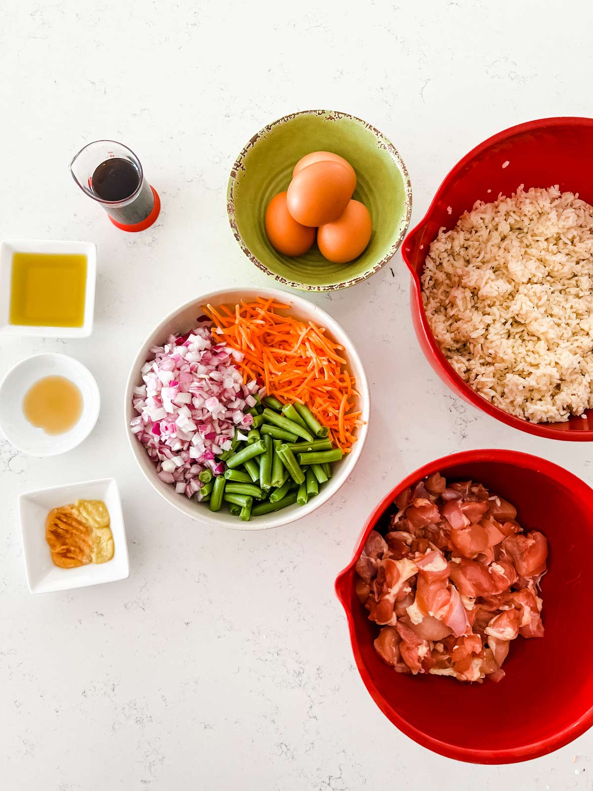 Overhead photo of cooked rice, cubed raw chicken, eggs, carrots, onion, green beans, garlic, ginger, soy sauce, oil and sesame oil.