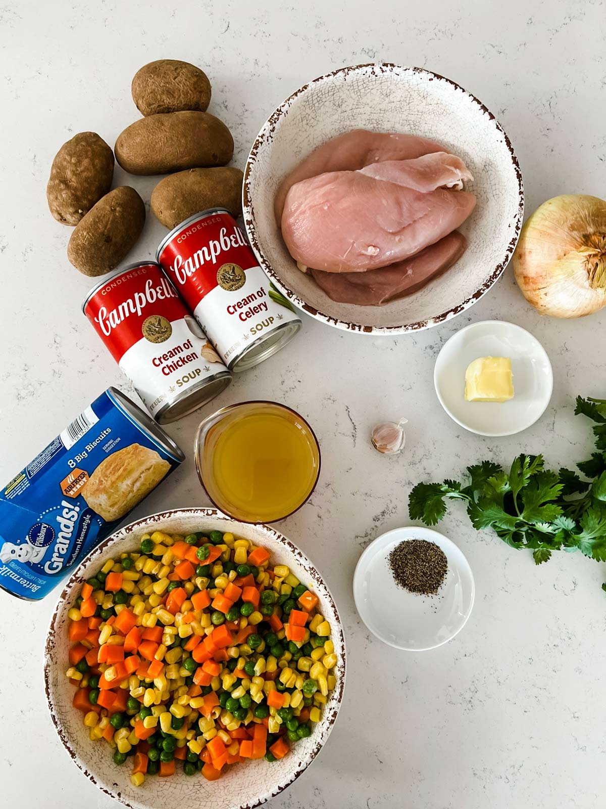 Overhead photo of chicken breast, mixed frozen vegetables, biscuits, cream of chicken soup, cream of celery soup, potatoes, broth, butter, onion, garlic, parsley , and seasonings.
