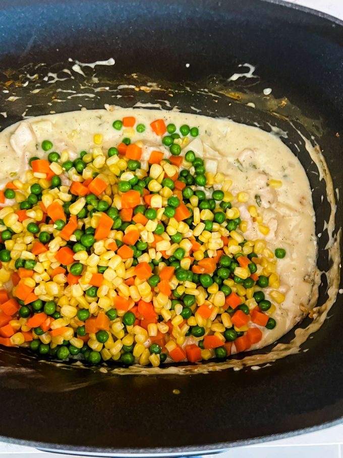 Peas, carrots, and corn being added to chicken pot pie filling in a slow cooker.