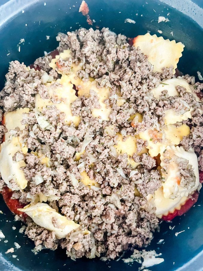 Crockpot ravioli that has been layered in the inner pot of a slow cooker with a top layer or ground beef.