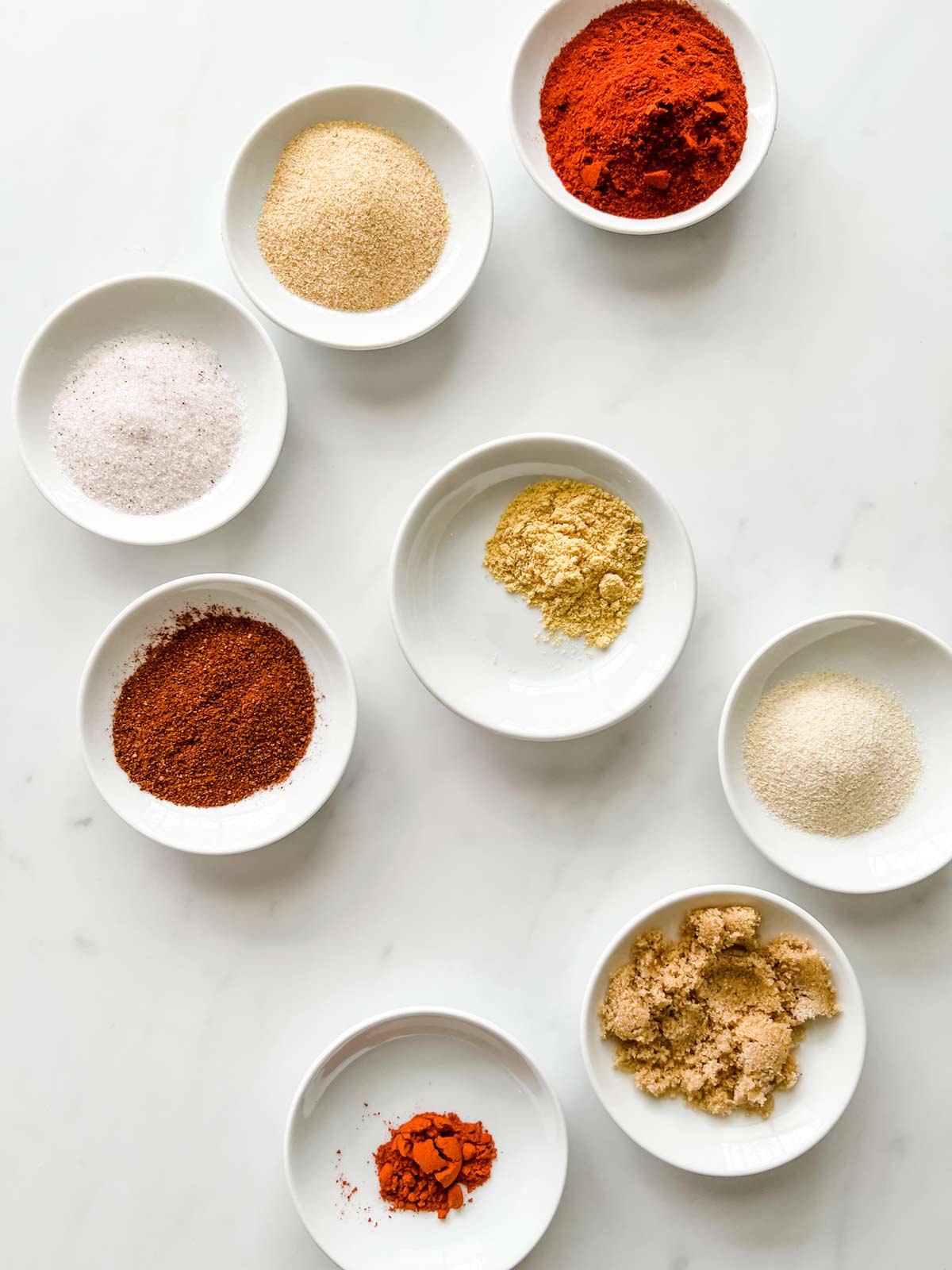 Overhead photo of small prep dishes of paprika, garlic powder, salt, brown sugar, onion powder, chili powder, ground mustard, and cayenne pepper.
