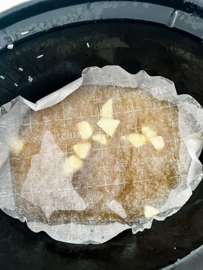 A slow cooker with white rice, broth, salt, and butter covered by a piece of parchment paper sitting directly on top of the rice.