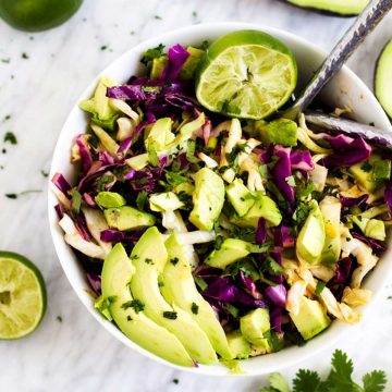 Square overhead photo of a white bowl with Cilantro Lime Coleslaw in it surrounded by avocado, cilantro, limes, and green onions.