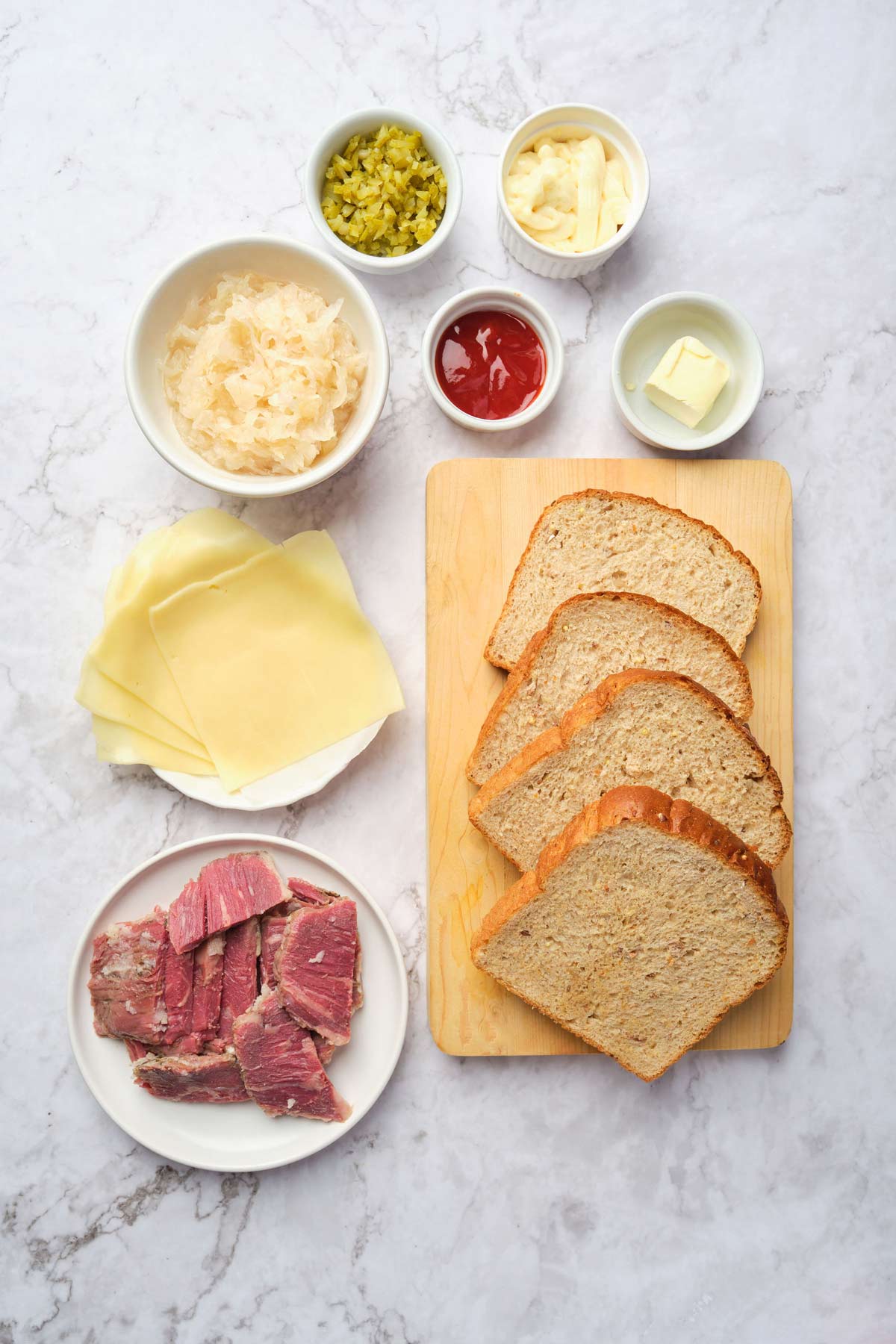 Overhead photo of rye bread, corned beef, swiss cheese, sauerkraut, pickles, ketchup, mayonnaise, and butter.