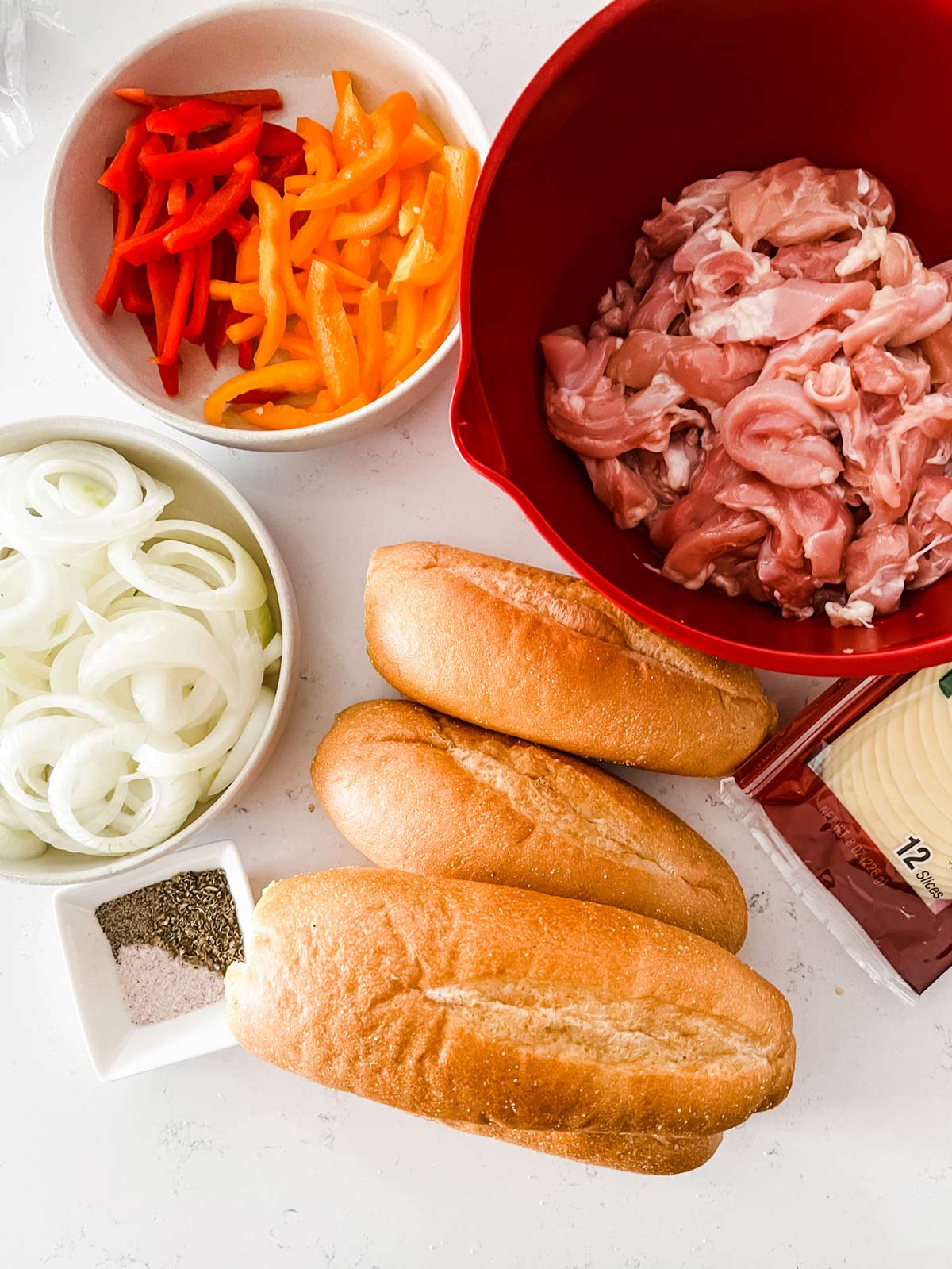 Overhead photo of sliced chicken thighs, red pepper, orange pepper, onion, seasonings and hoagie buns on a white countertop.