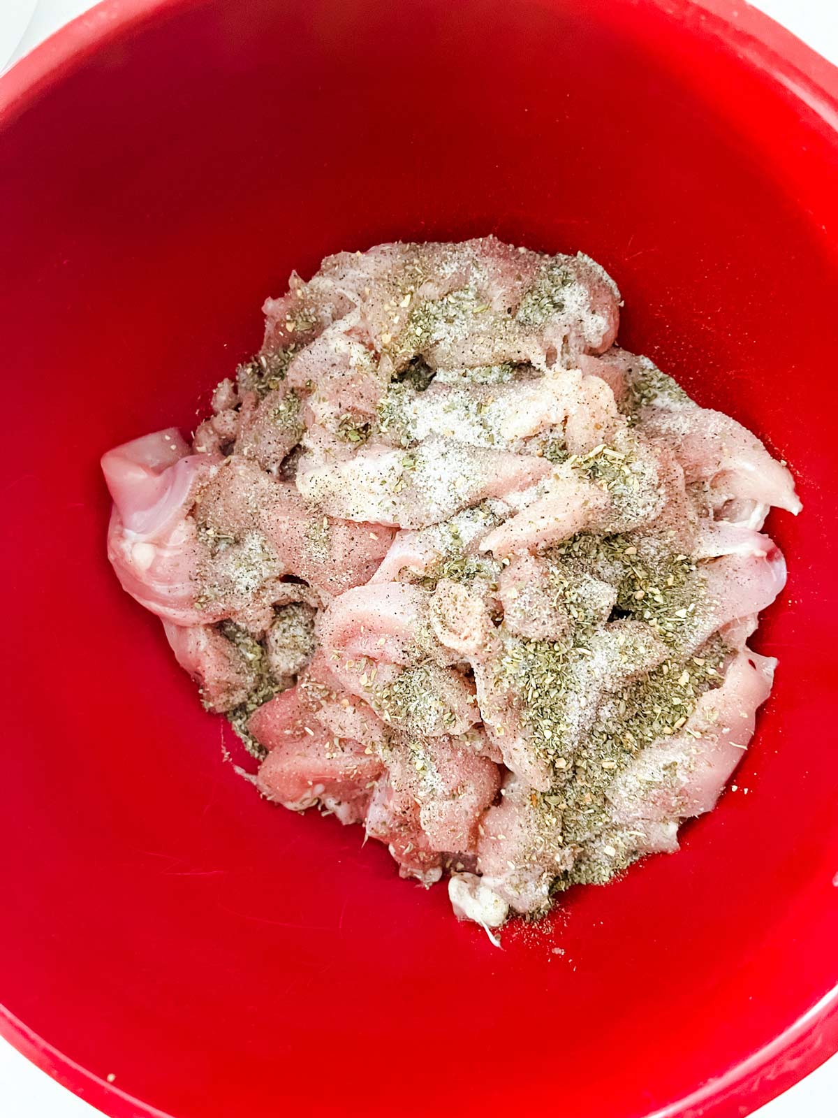 Sliced chicken thighs in a red bowl with seasonings on top.