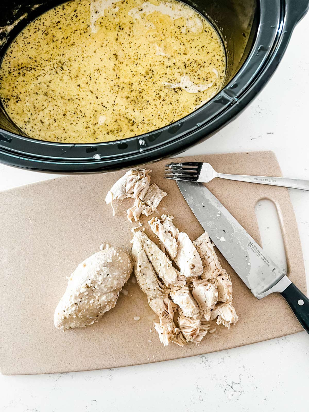 Chicken being cut o a cutting board next to a slow cooker with creamy sauce.