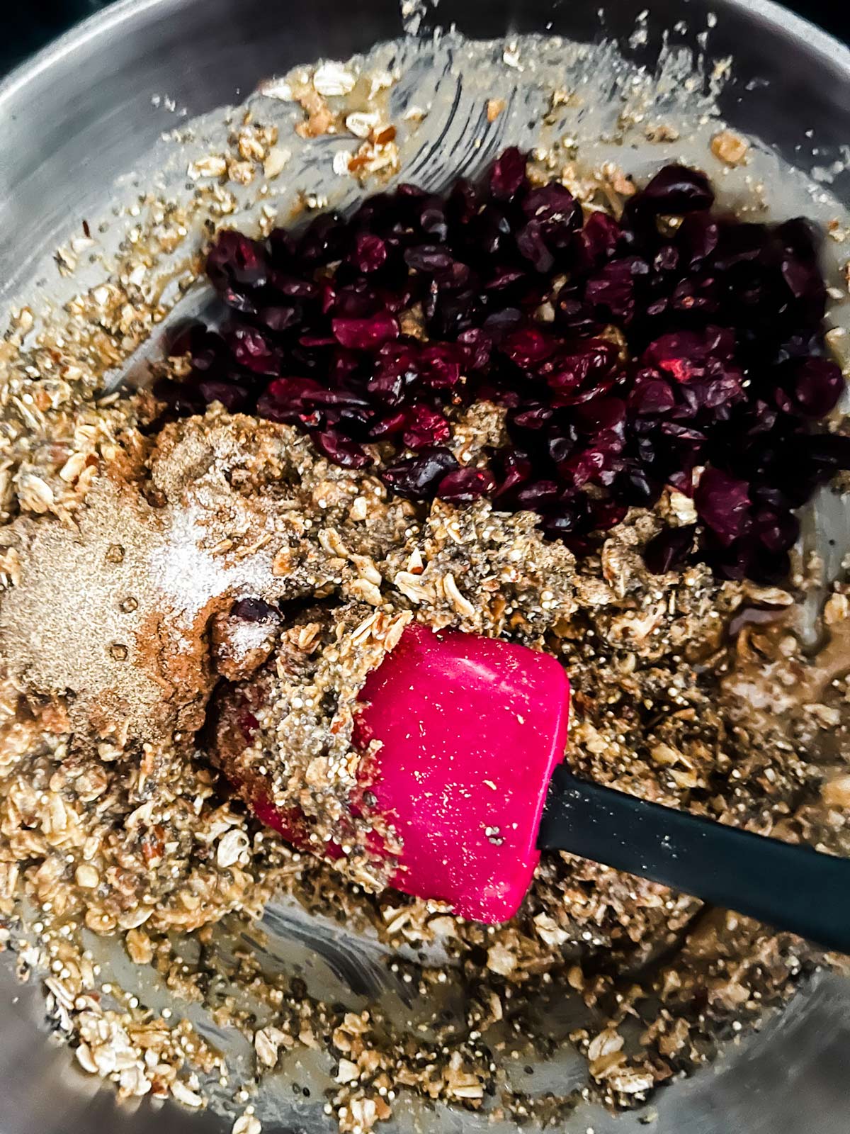 Honey, peanut butter, Quinoa, chia seeds, flax seeds, shredded coconut, oatmeal, and almonds that have just had cranberries added to it and are being stirred with a spatula.