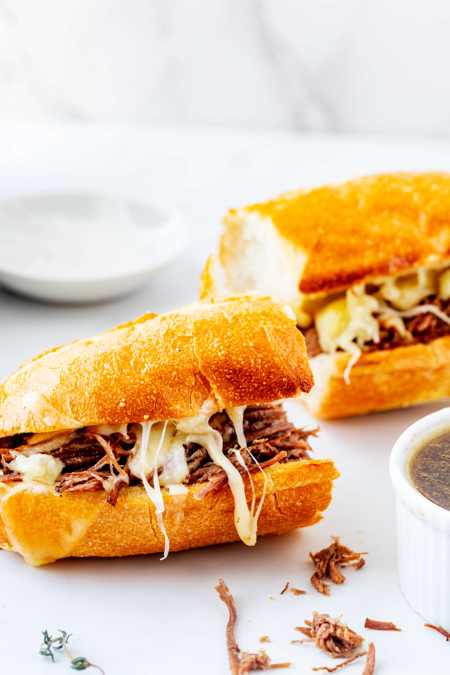 Photo of a slow cooker french dip sandwich with cheese sitting on a white counter with a ramekin of au jus next to it.
