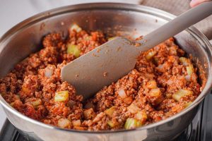 Tomato sauce and tomato paste being stirred into a skillet of ground beef.