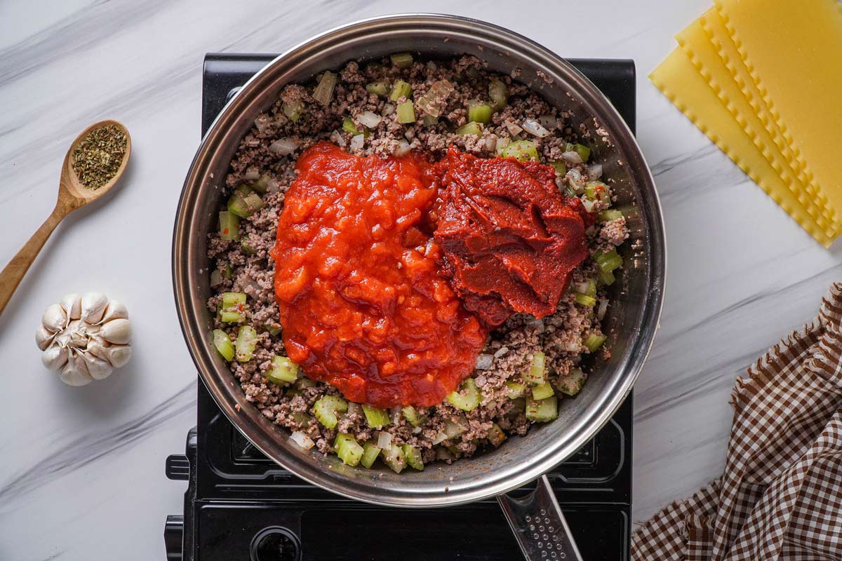Crushed tomatoes and tomato paste being added to a skillet of seasoned ground beef and vegetables.