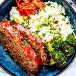 Square overhead photo of crockpot meatloaf plated over a bed of mashed potatoes with roasted tomatoes and broccoli.
