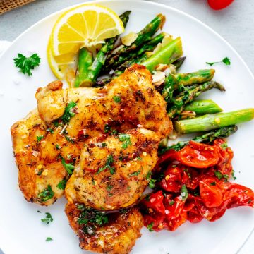Square overhead photo of Ninja Foodi Chicken Thighs on a white plate with roasted tomatoes, asparagus, and a lemon wedge.