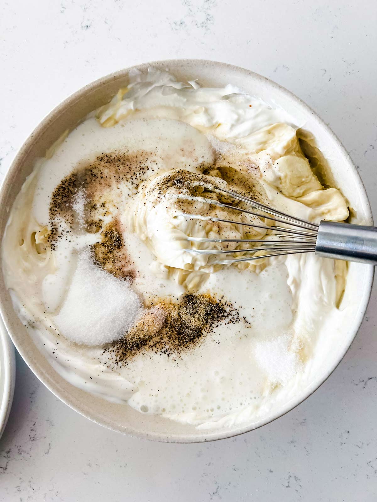 Photo of mayo, sour cream, cream and seasonings that have been dumped into a bowl with a whisk.