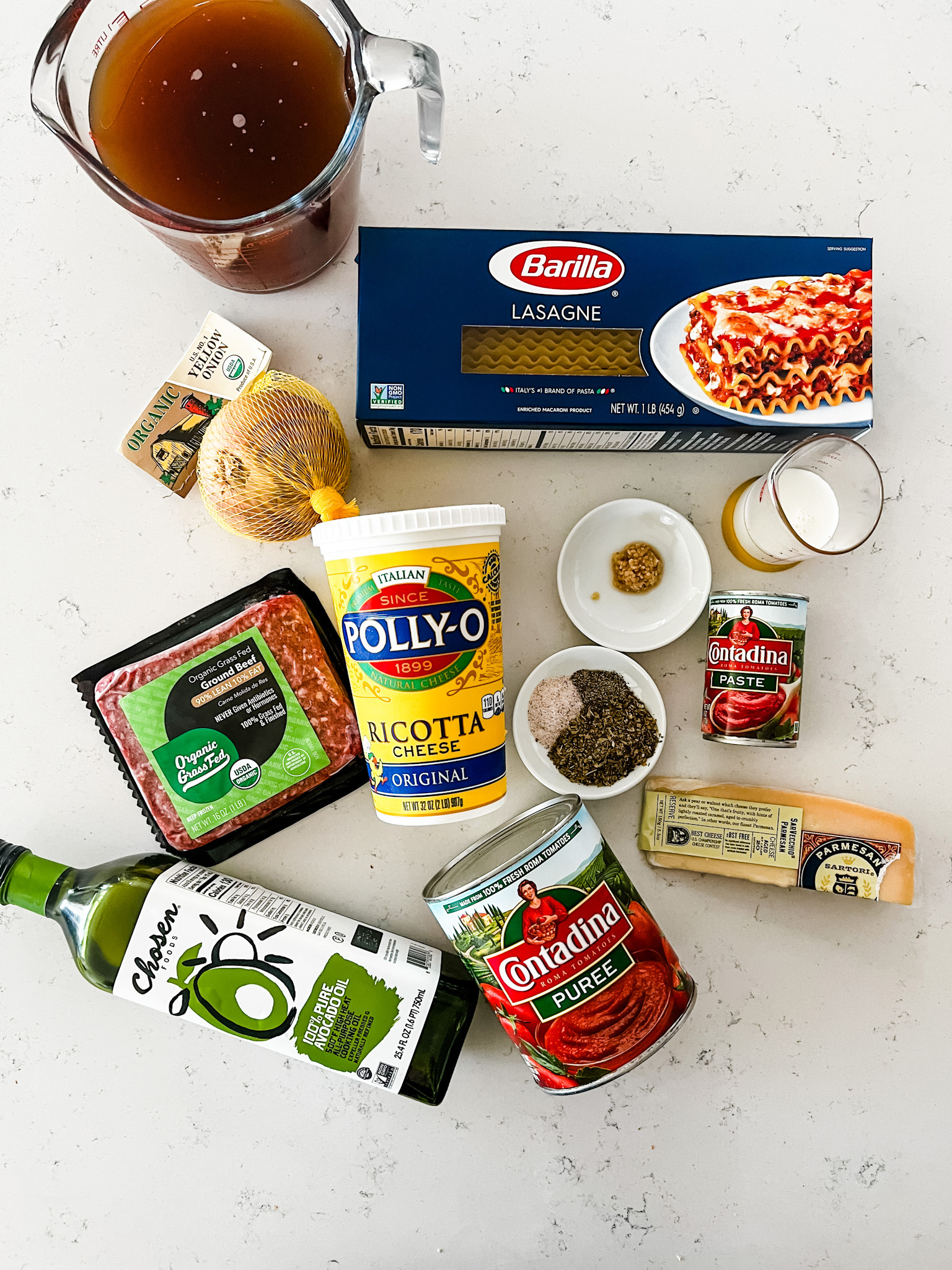 Overhead photo of a package of lasagna noodles, beef broth, ricotta, ground beef, oil, onion, garlic, parmesan, tomato puree, tomato paste, and seasonings.