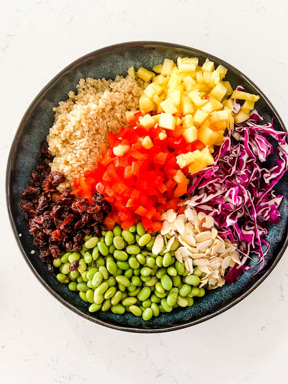 A blue bowl with quinoa, edamame, pineapple, red pepper, dried cranberries, cabbage, and almonds.