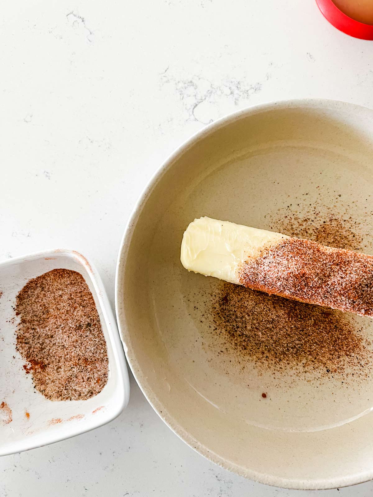A small bowl of seasonings that has had half of its contents sprinkled on top of butter.