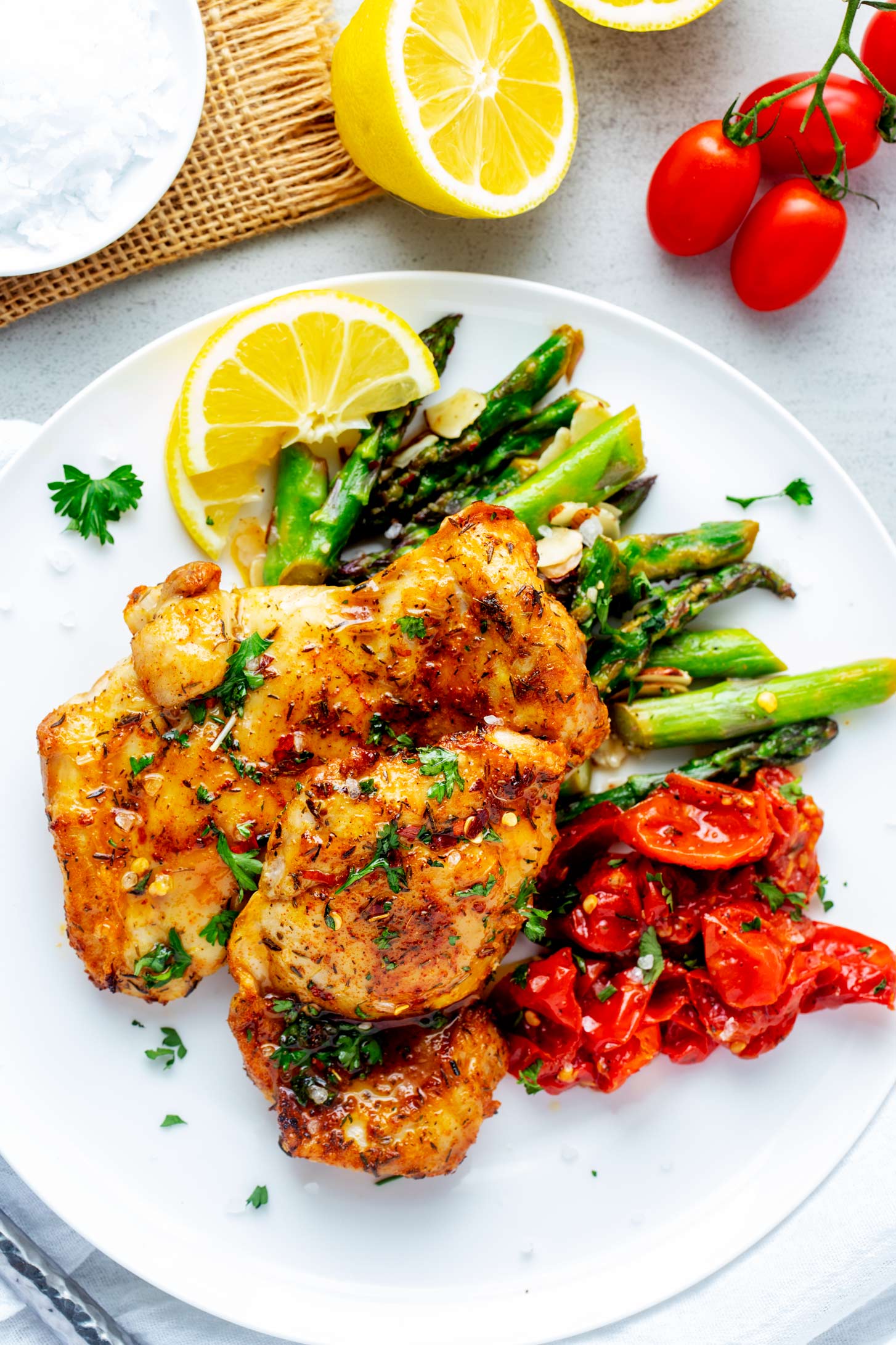 Overhead photo of Ninja Foodi Chicken Thighs on a white plate with roasted tomatoes, asparagus, and a lemon wedge.