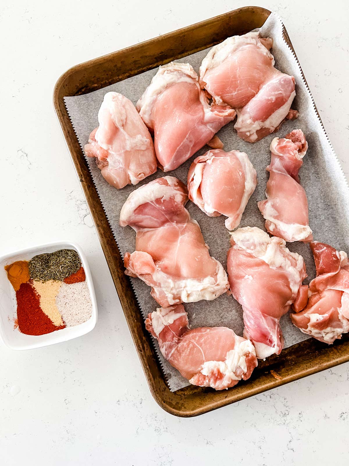 Overhead photo of a parchment lined tray with raw chicken thighs sitting next to a small dish of seasonings.