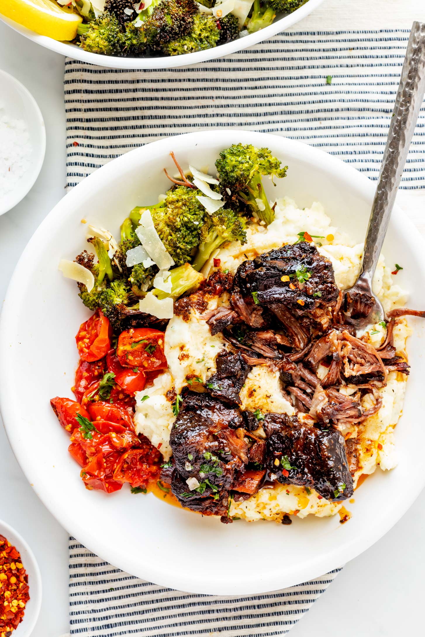 Overhead photo of slow cooker short ribs over mashed potatoes on a plate with roasted tomatoes and broccoli.