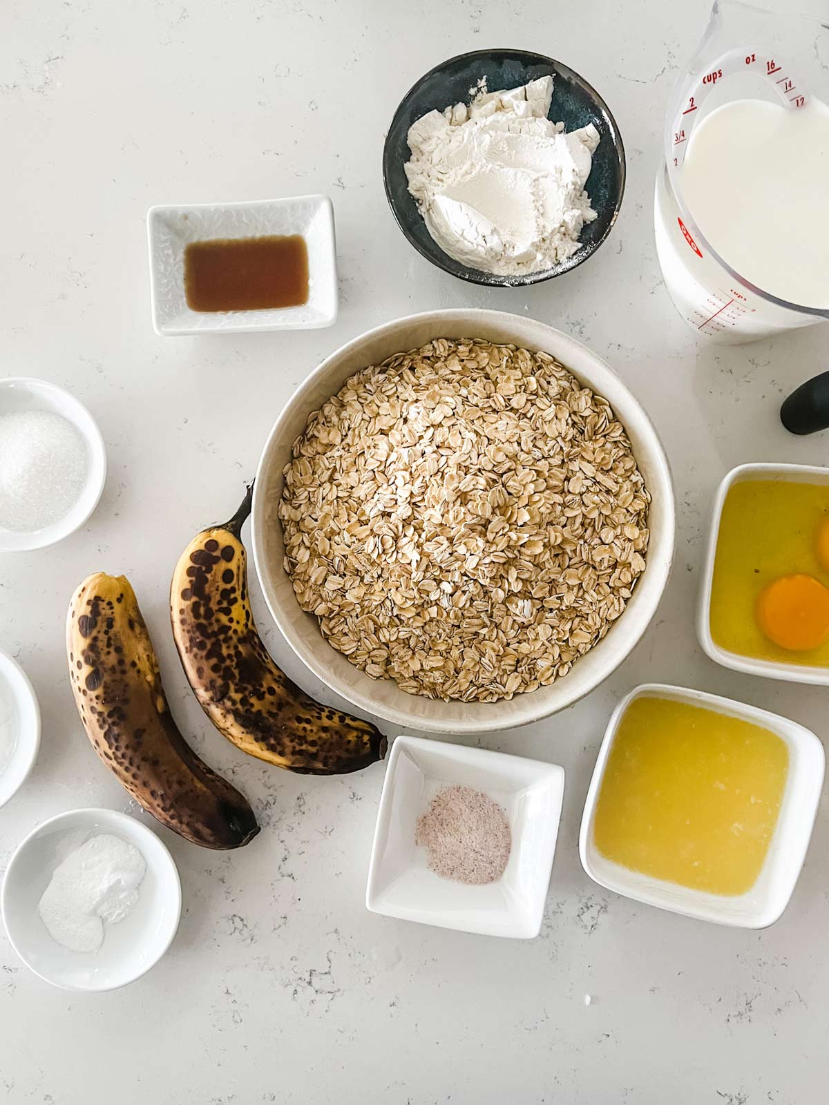Overhead photo of oats, flour, sugar, baking powder, baking soda, salt, milk, eggs, bananas, butter, and vanilla extract.