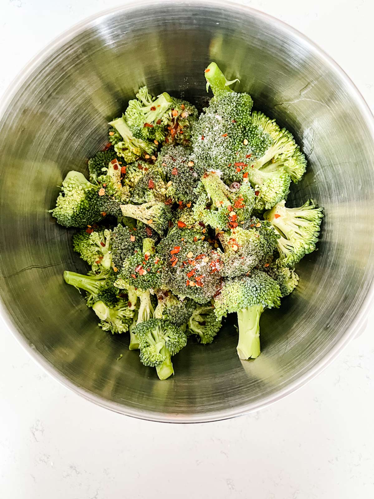 Broccoli florets in a metal bowl with seasonings on top of them.