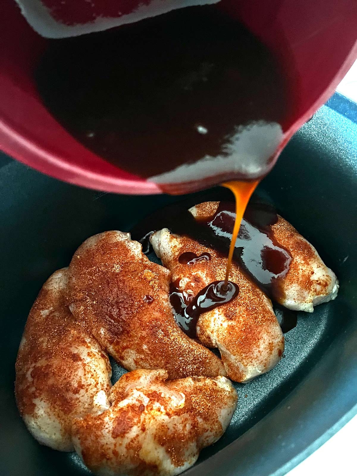 BBQ sauce being poured over chicken in a slow cooker.