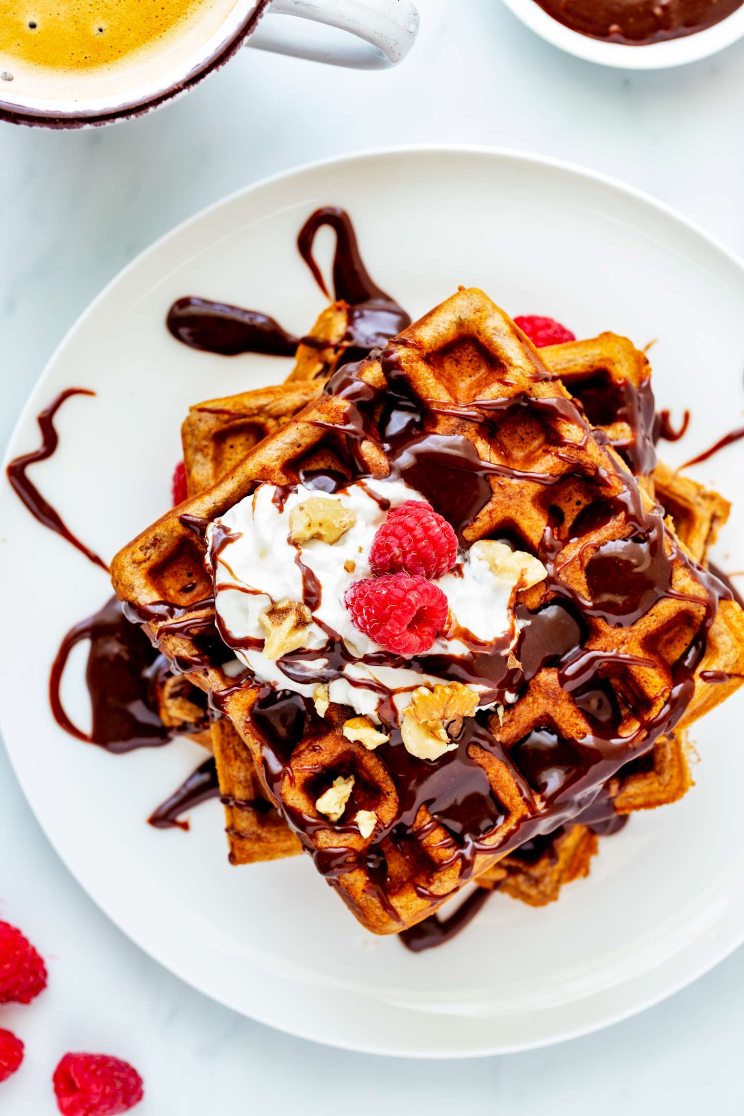Overhead photo of nutella waffles on a white plate with nutella, whipped cream, berries, and nuts.