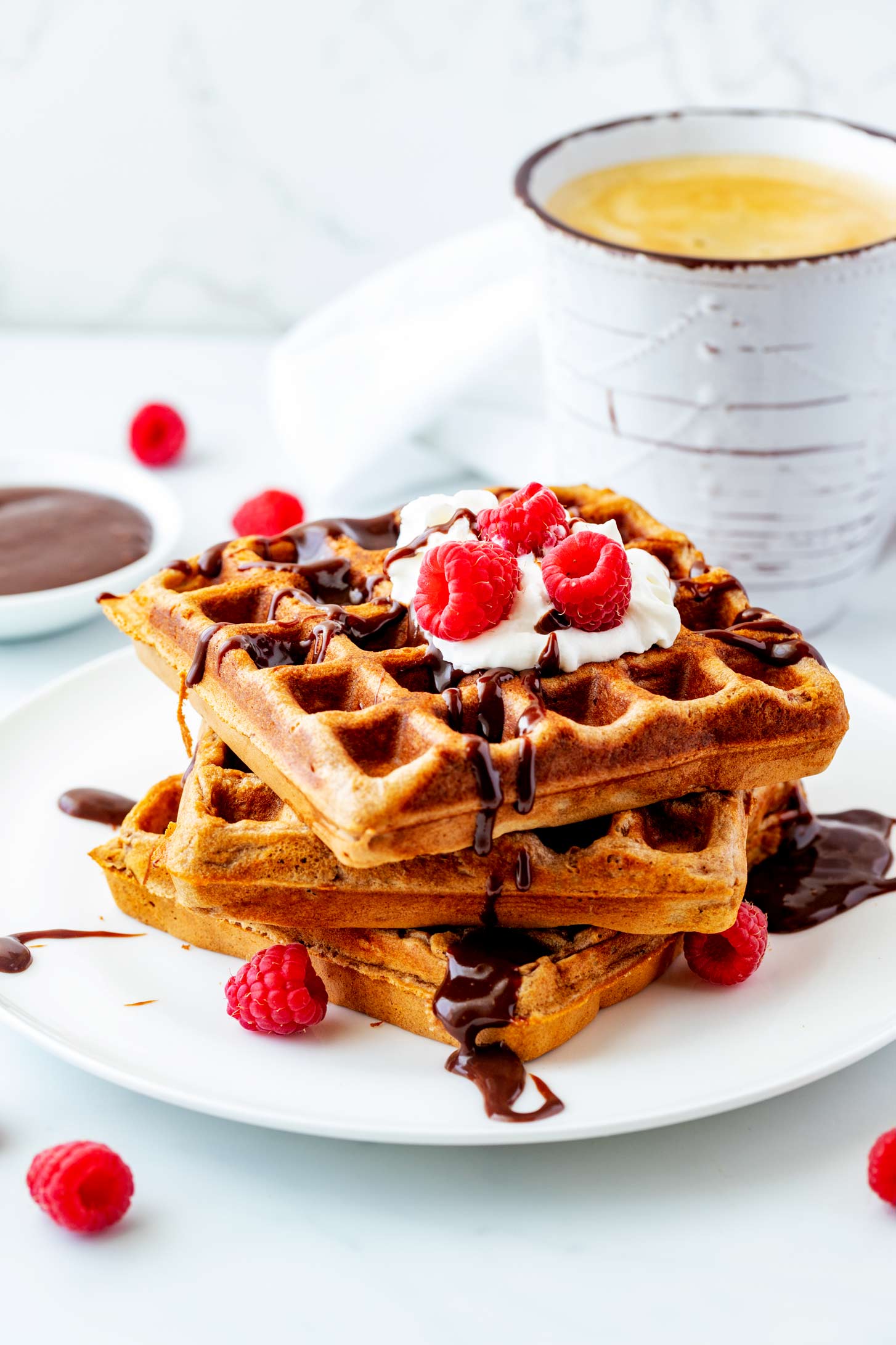 Side photo of a white plate of stacked nutella waffles garnished with nutella, whipped cream and raspberries.